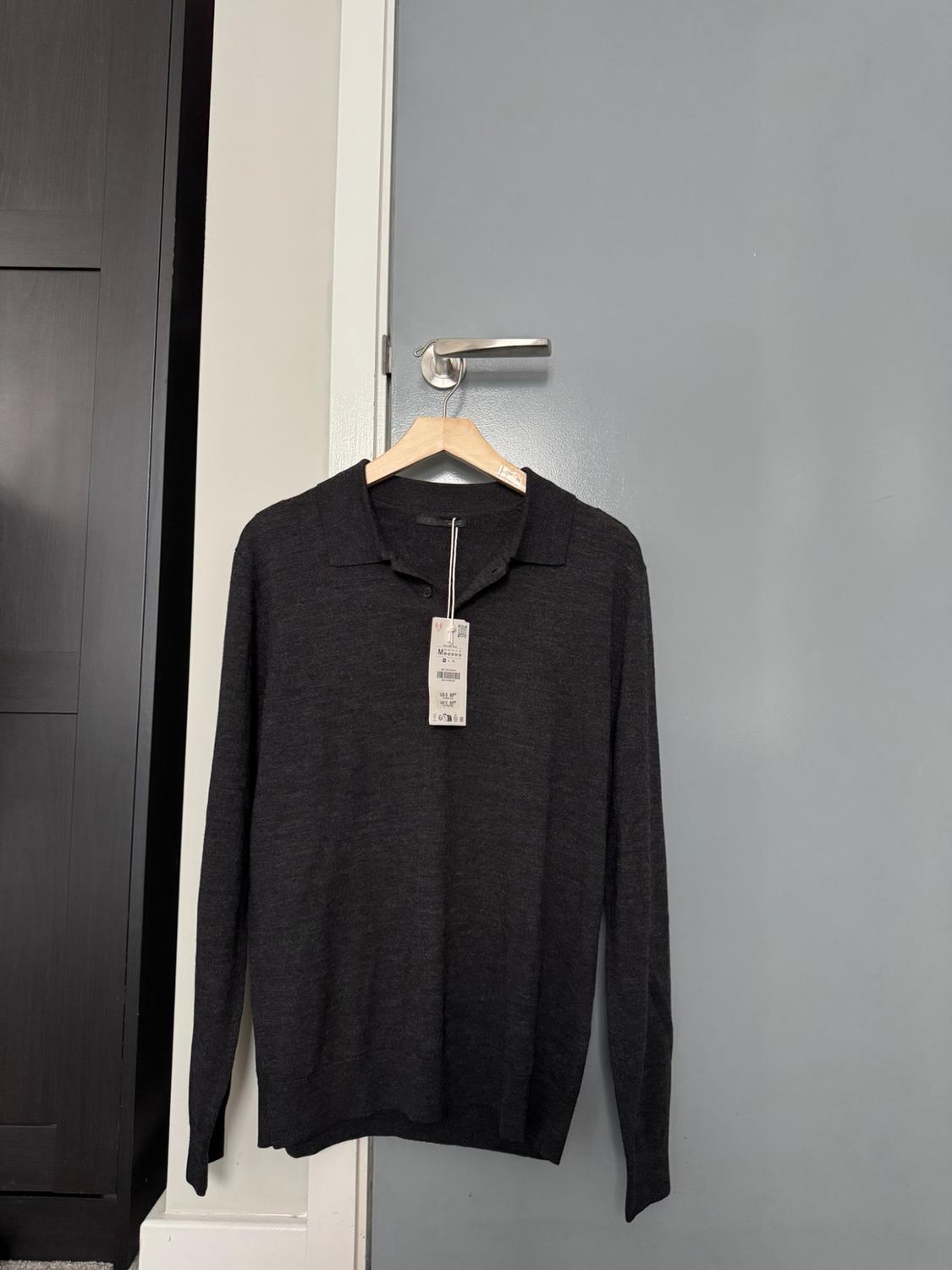 Dark grey long-sleeved polo shirt with a label, hanging on a wooden hanger over the metal knob of a blue-grey door.