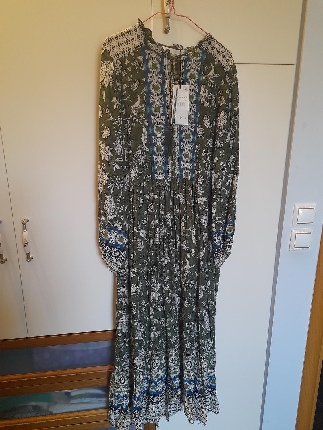 Long dress hanging on a hanger, with a floral print in green and blue tones, and lace details on the collar and sleeves.