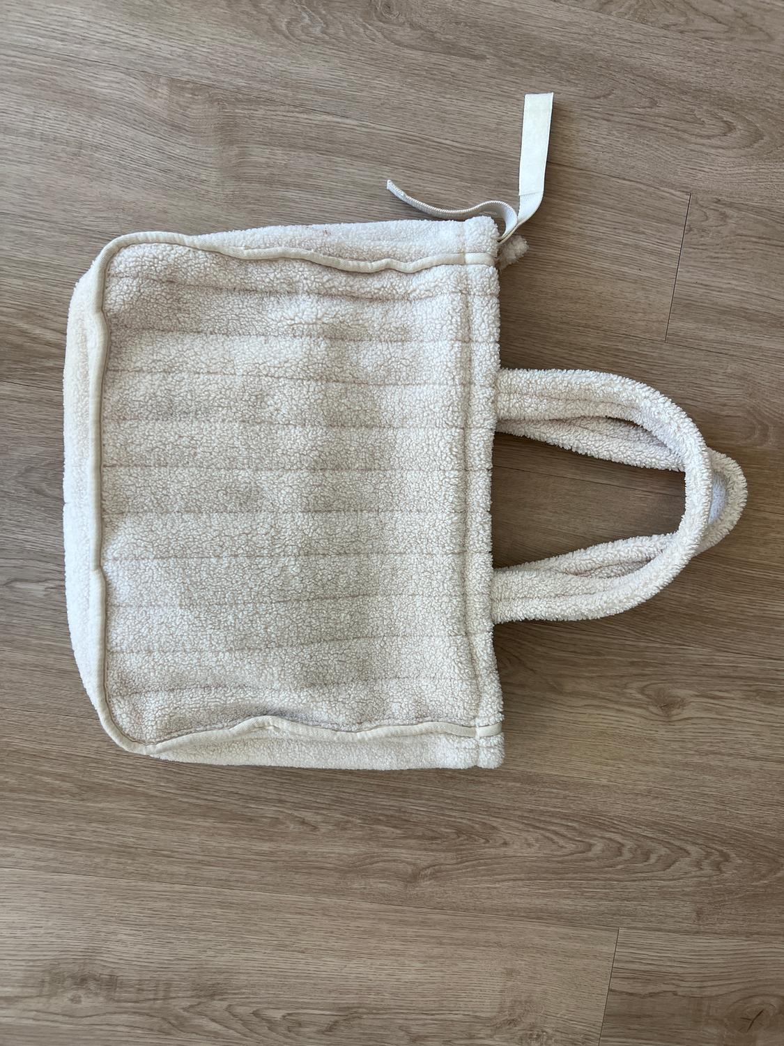 Soft, fluffy cream fabric bag on a light wooden floor. It has thick handles and a white tag.