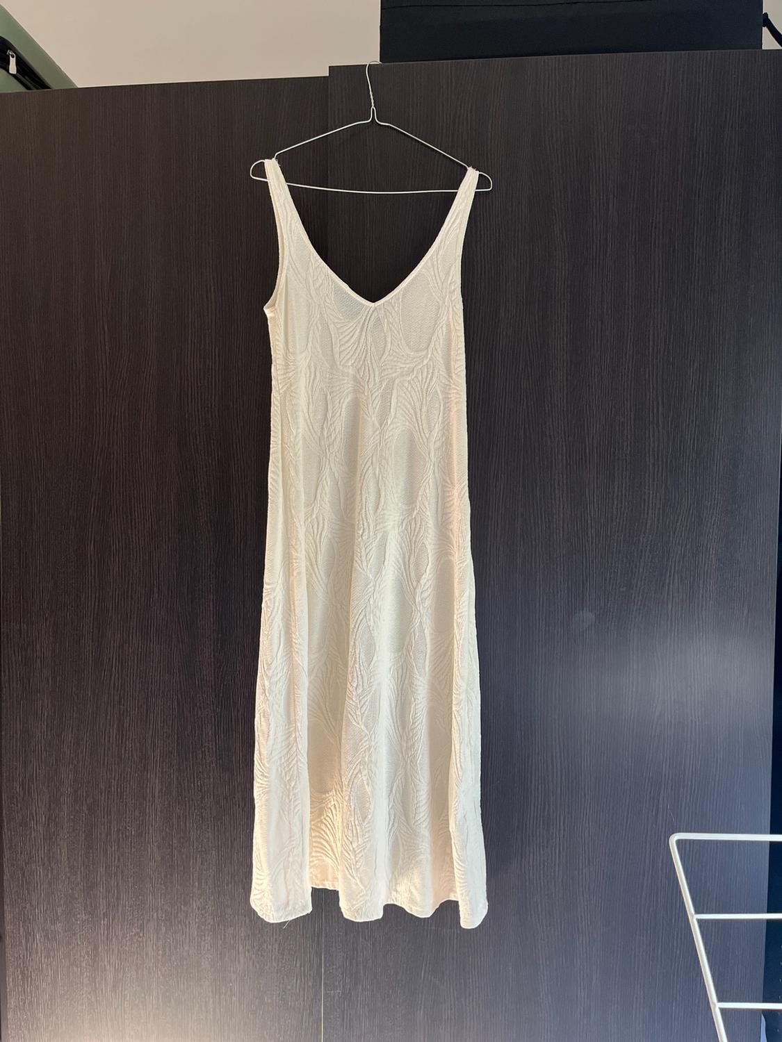 Long white sleeveless dress with a V-neck and leaf texture, hanging on a hanger in front of a dark wooden wardrobe.