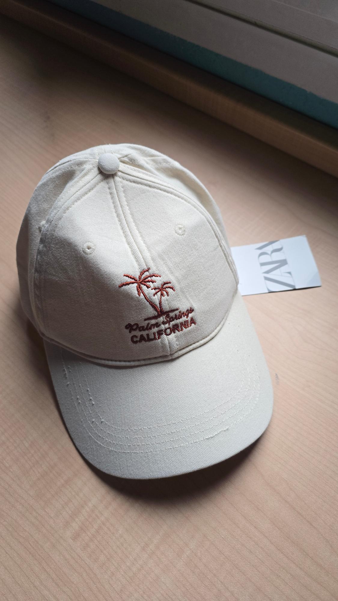 White cap with palm tree embroidery and red "Palm Springs CALIFORNIA" text. ZARA label in the background on a wooden table.