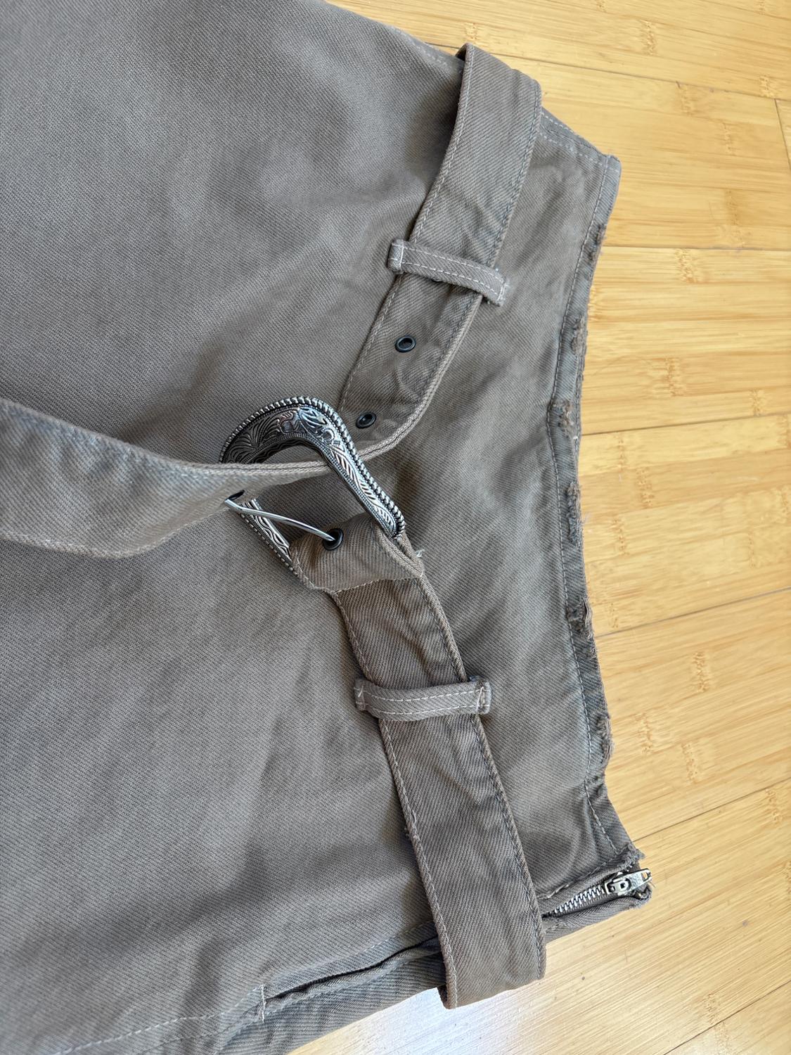 Brown thick fabric pants with a belt of the same material and decorative metal buckle on a light wood floor.