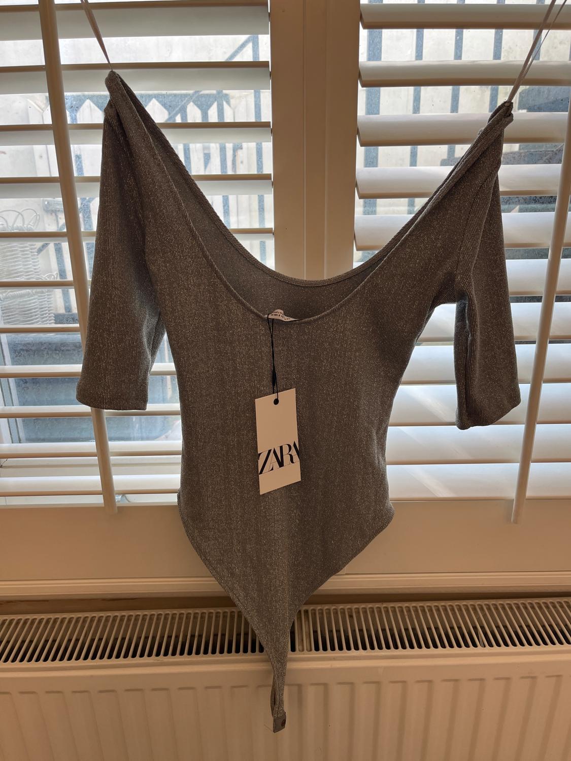 Grey long-sleeved babygrow hanging in front of a window with white shutters. Label visible with the text "ZARA". Indoor setting.