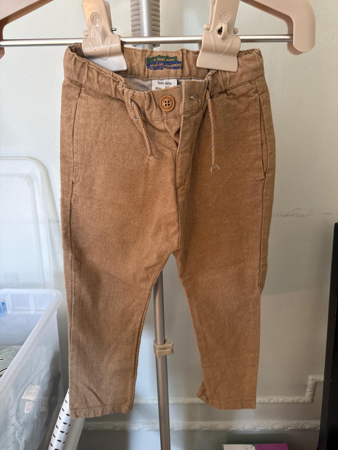 Brown baby trousers hanging on a hanger, with drawstrings and a front button. Visible label: "Zara baby size 6-9 months".