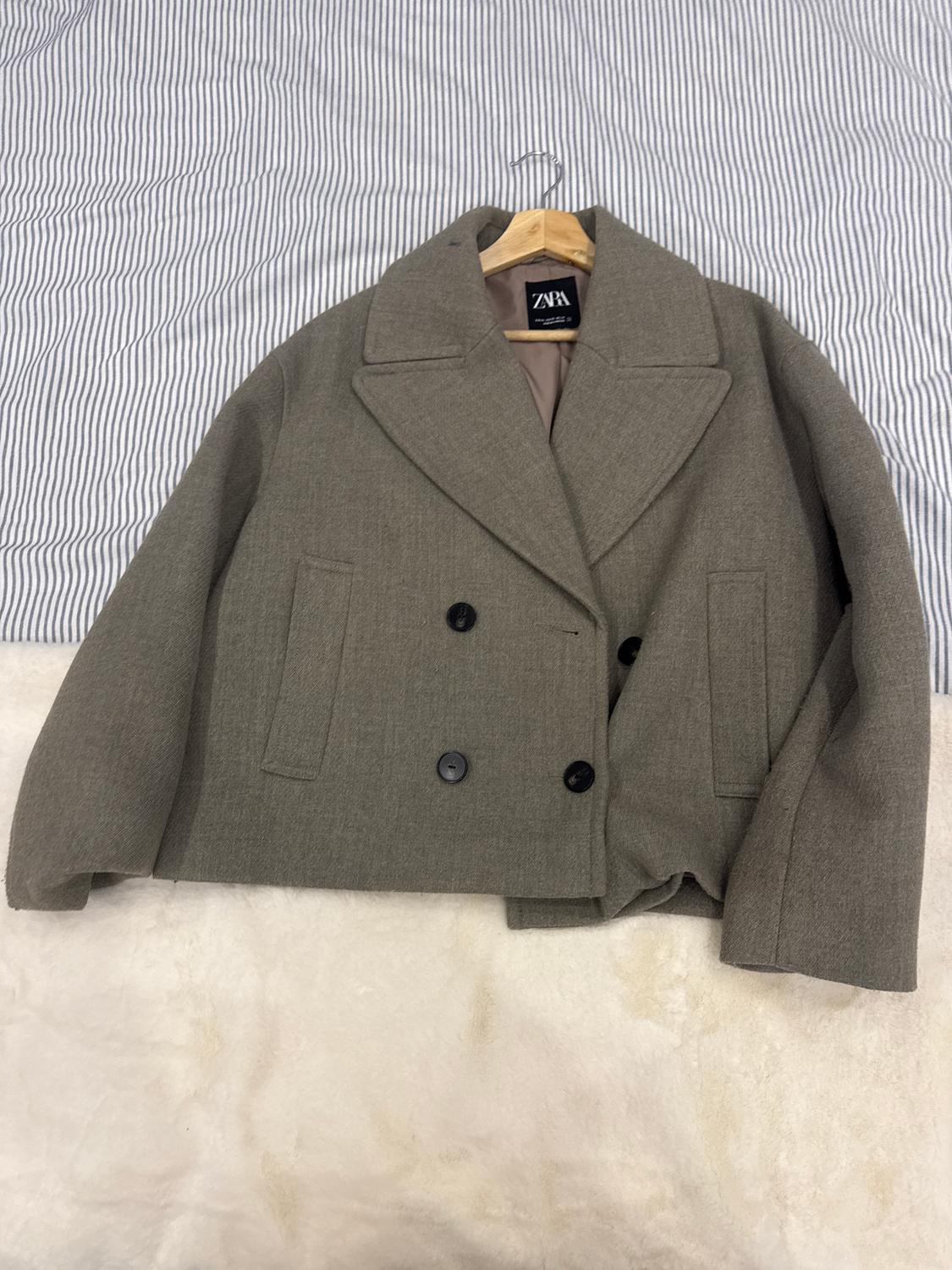 Grey double-breasted coat hanging on a hanger over striped fabric. Visible label with the text "ZARA".