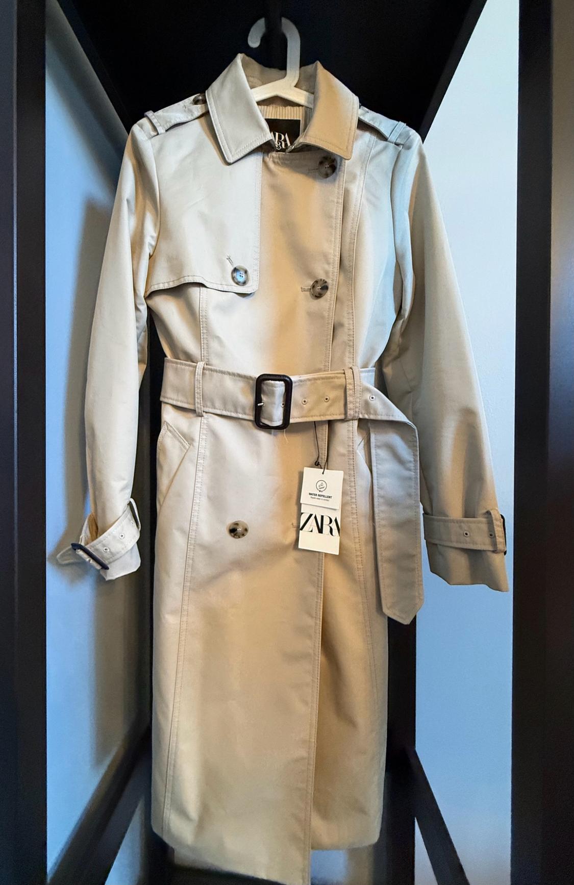 Beige gabardine by zara hanging on a hanger. Features a belt with a buckle, buttons, and brand tags from zara.