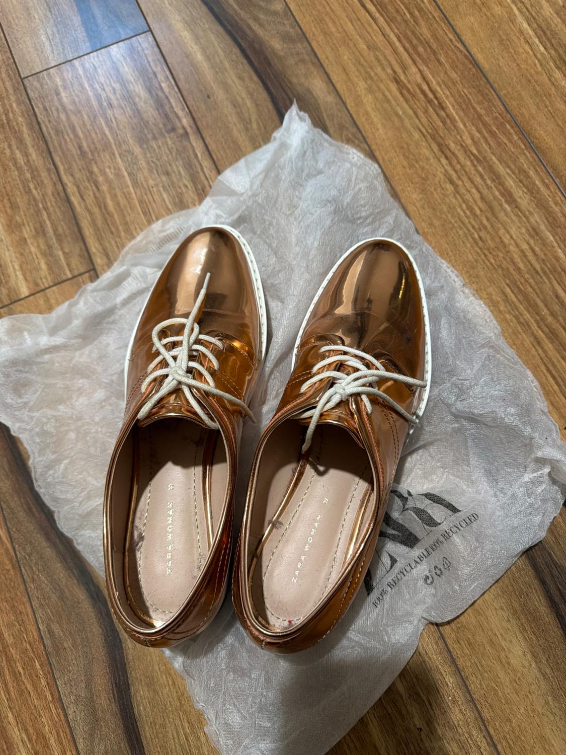 Copper metallic shoes with white laces on a bag on a wooden floor. Text: ZARA WOMAN 39, 100% RECYCLABLE 100% RECYCLED.
