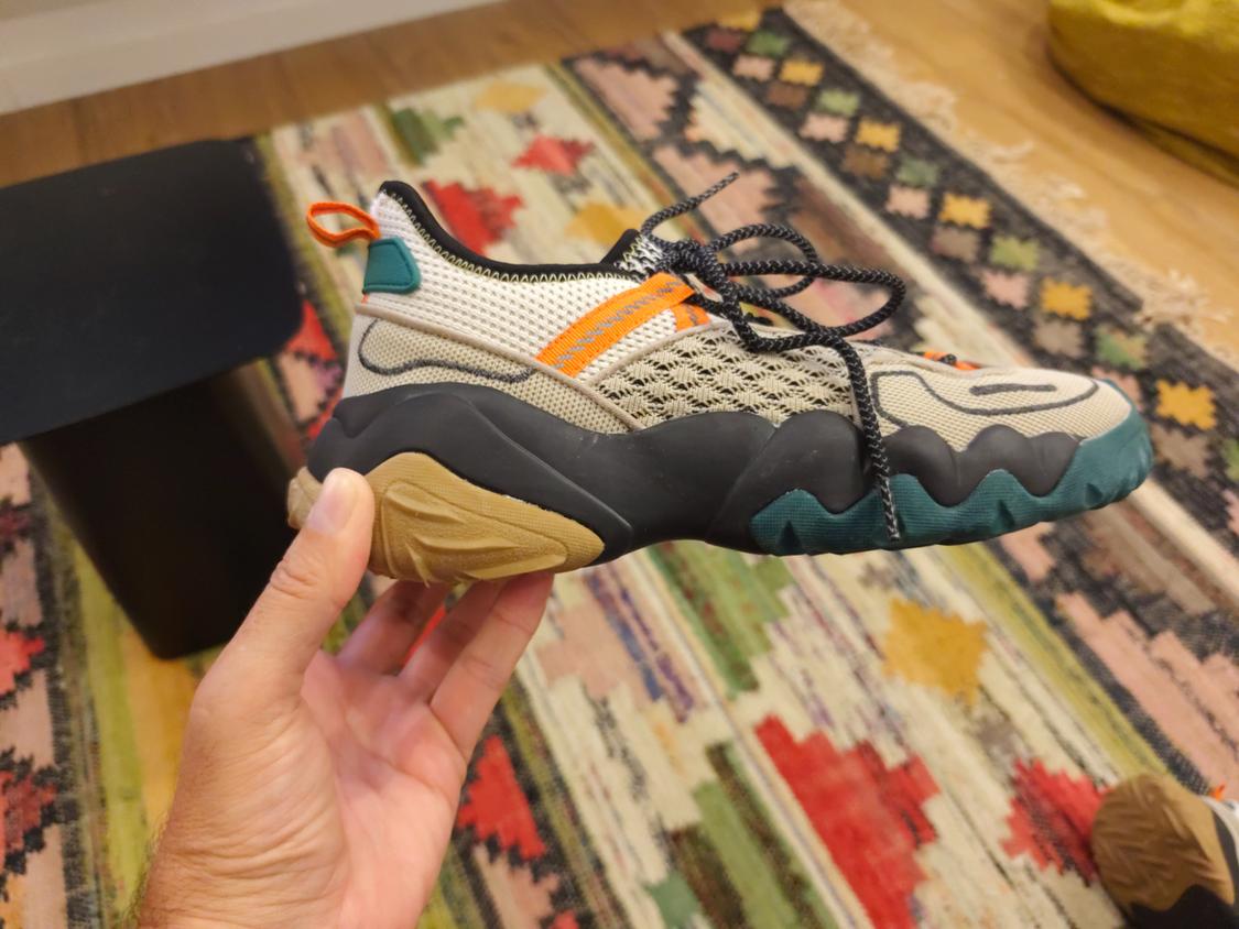 Hand holding a beige trainer with orange details and a thick black sole, above a colourful geometric rug.