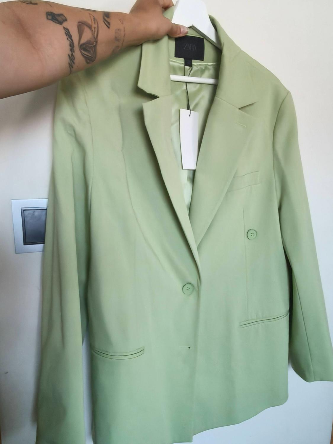 A tattooed arm holds a light green blazer with a ZARA label on a white hanger in front of a wall with a light switch.