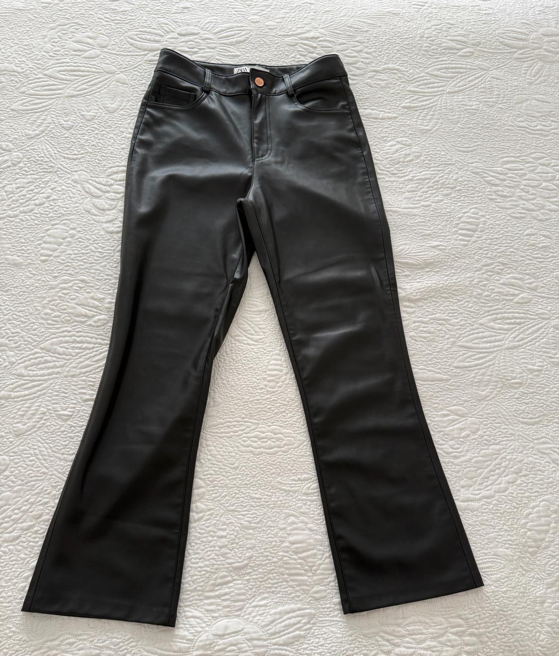 Black trousers with a glossy finish and flared cut resting on a textured white bedspread. ZARA label visible on the waistband.