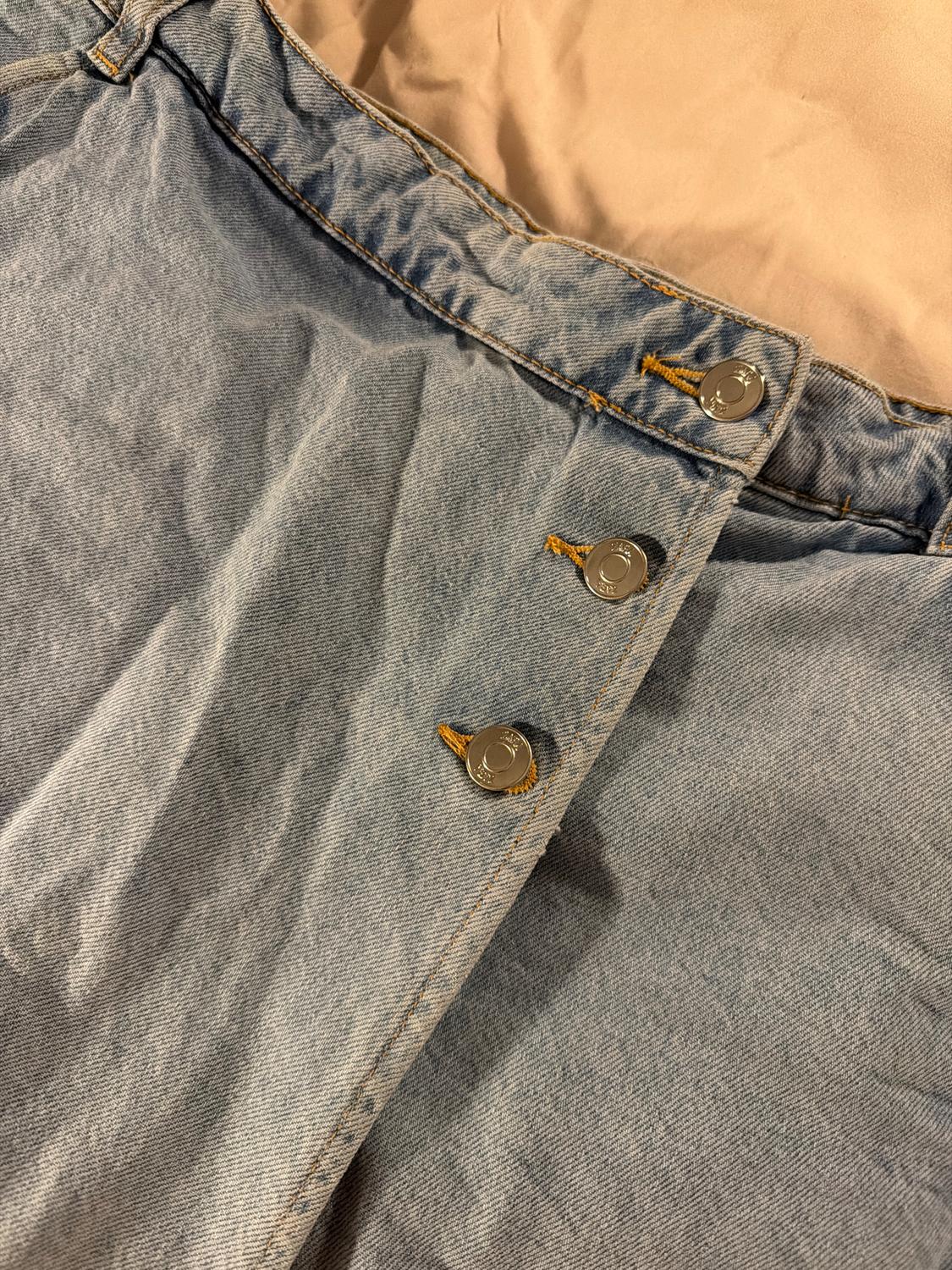 Detail of a light blue denim skirt with metallic buttons and visible stitching on a beige surface.