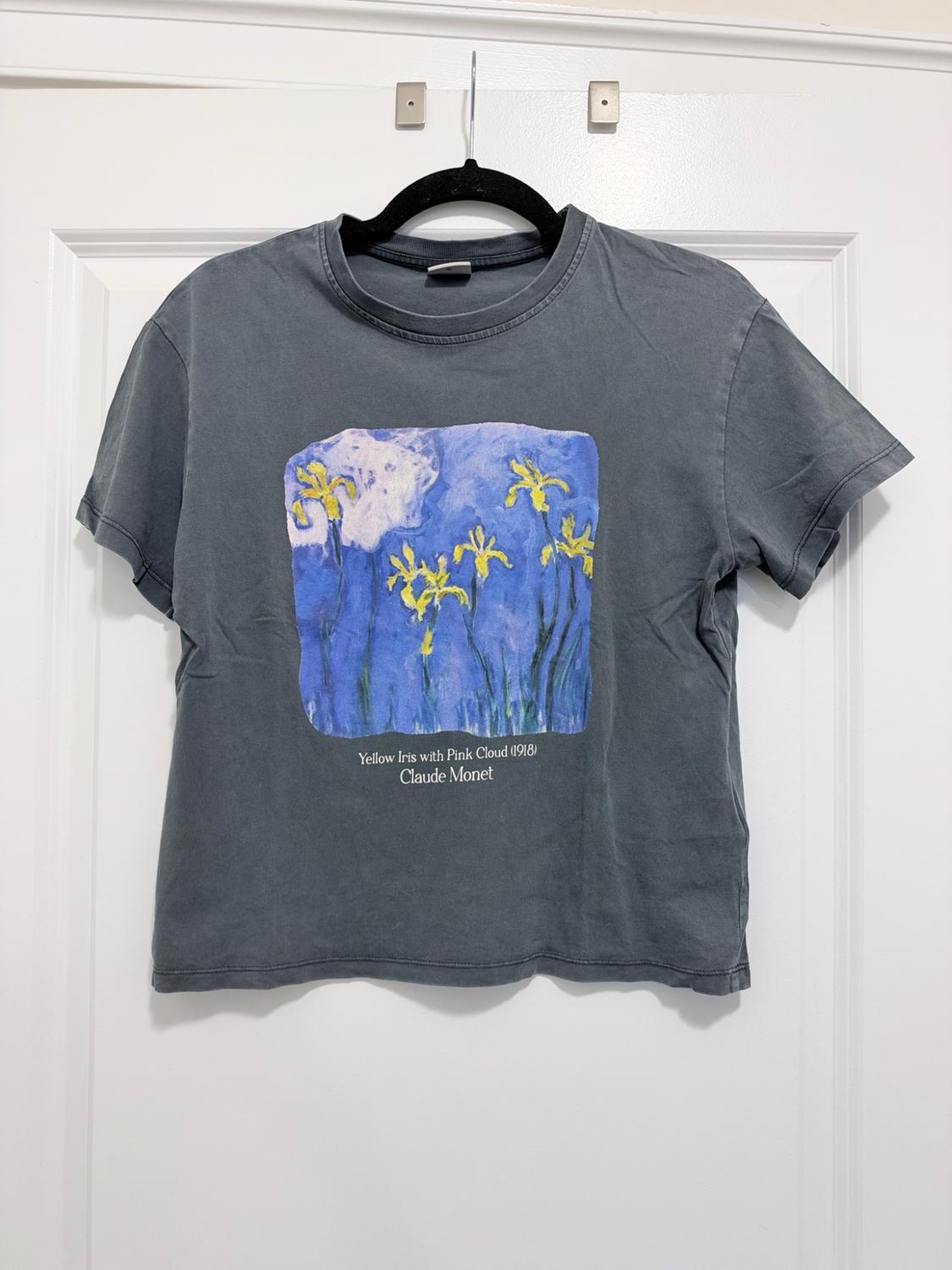 Dark gray t-shirt hanging on a white door with print and text: Yellow Iris with Pink Cloud (1918) Claude Monet.