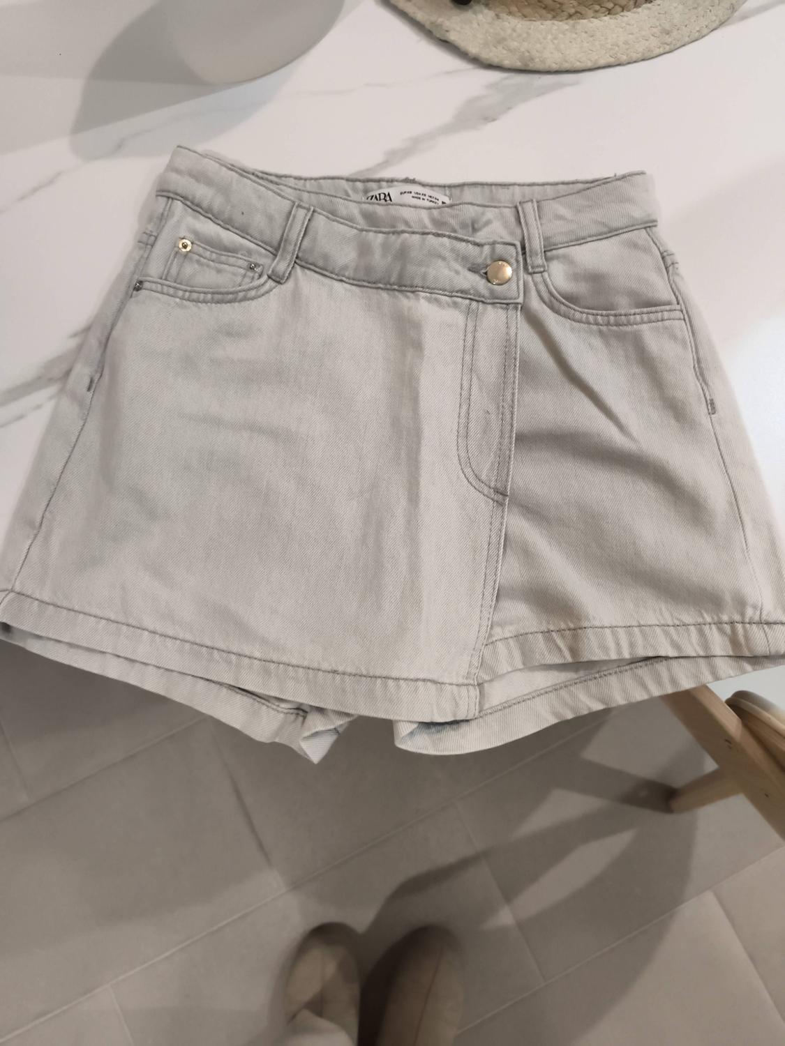 Light grey denim shorts on a white table. Stitching details and metal buttons are visible. Part of a hat in the background.