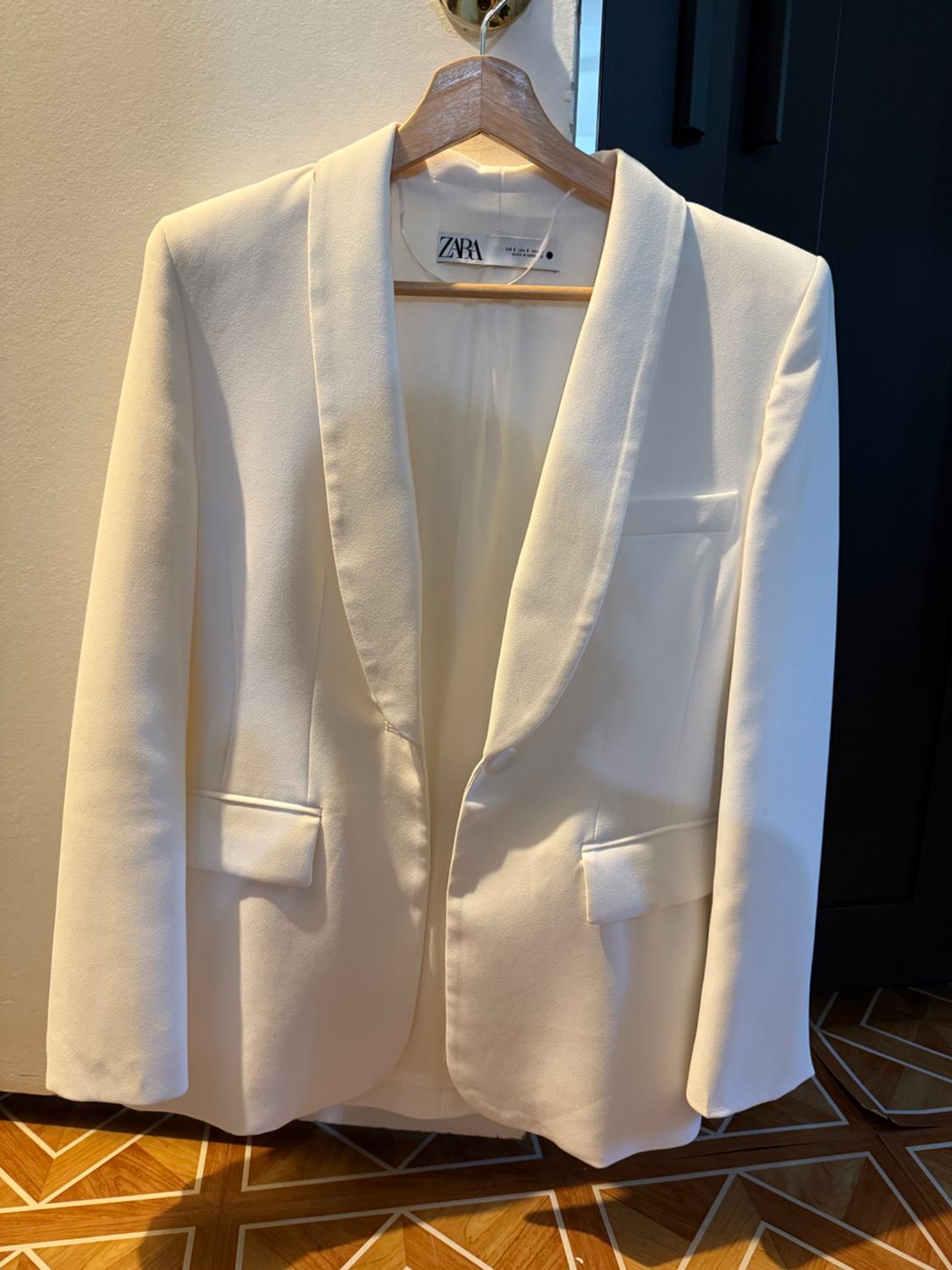 White Zara jacket hanging on a wooden hanger, with wide lapels and pockets, against a light wall background and wooden floor.