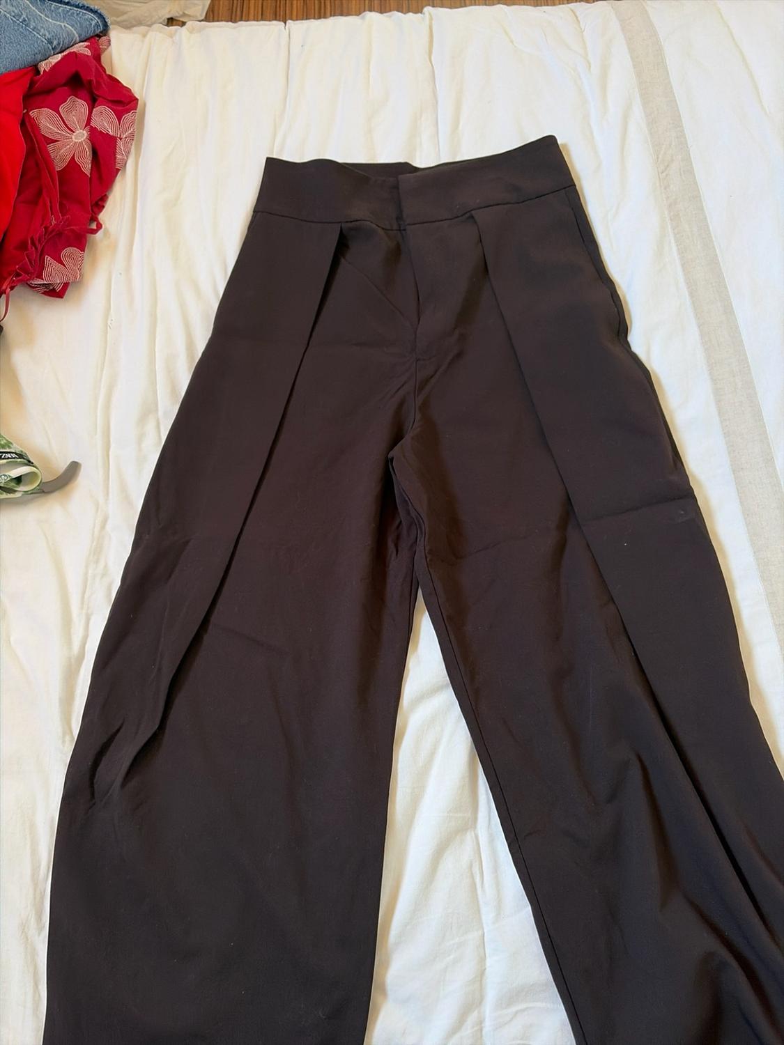 Dark brown wide-leg trousers with front pleats, laid out on a crumpled white sheet next to other garments.