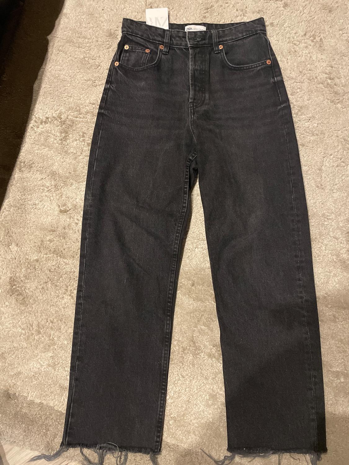 Black straight-cut jeans with a frayed hem, on a beige rug. Visible label on the waistband.