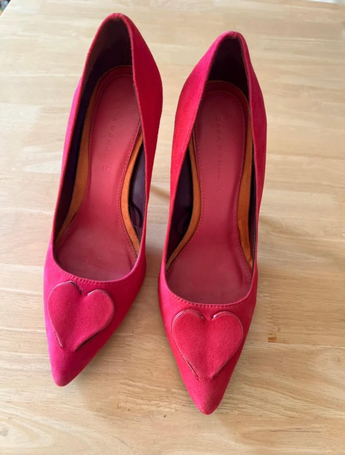 Red high-heeled shoes with heart detail on the toe, on a light wooden surface. Visible text: "ZARA WOMAN".
