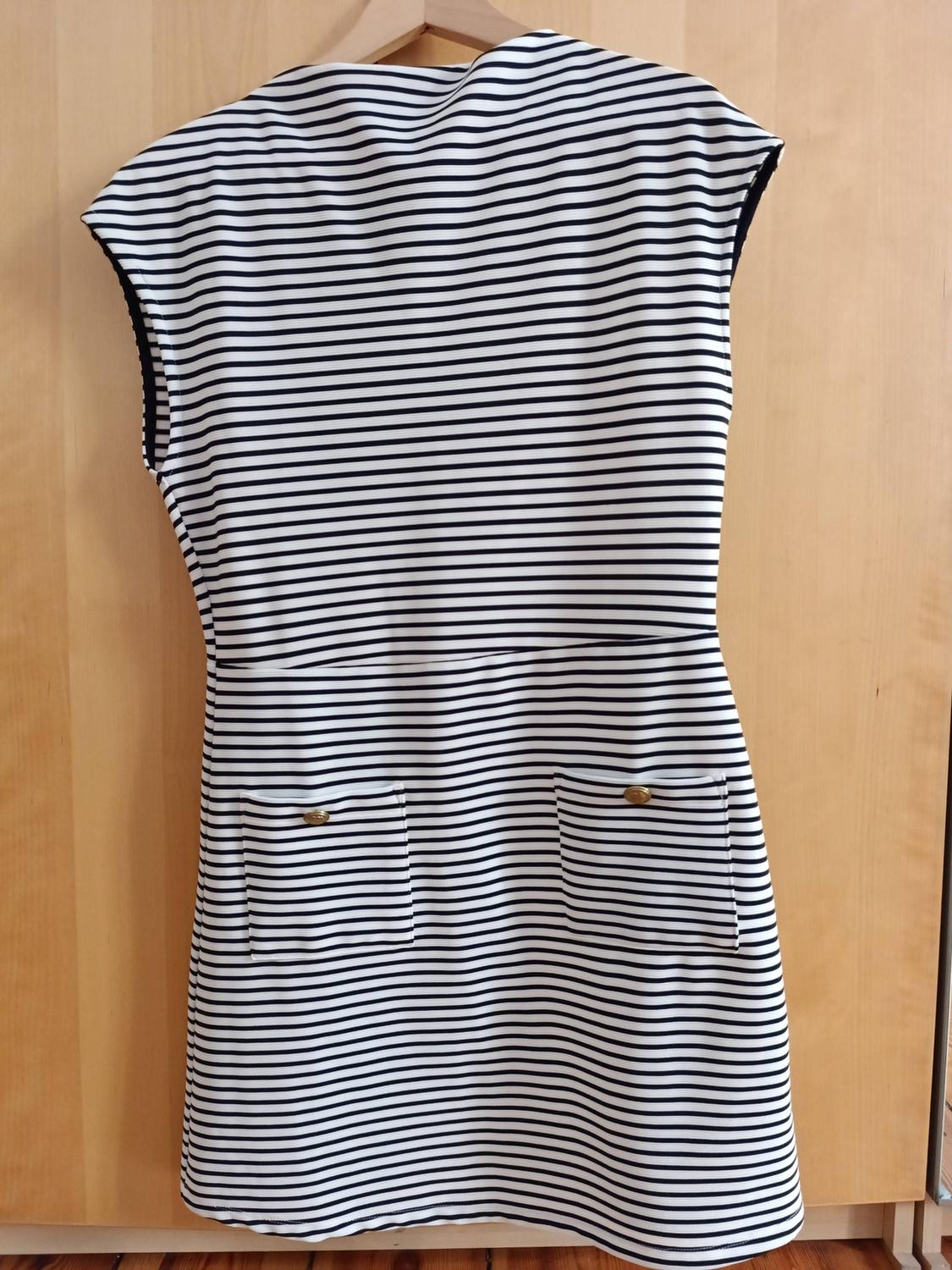 Short black and white horizontal striped sleeveless dress, with gold button pockets, hanging on a hanger against a wooden background.