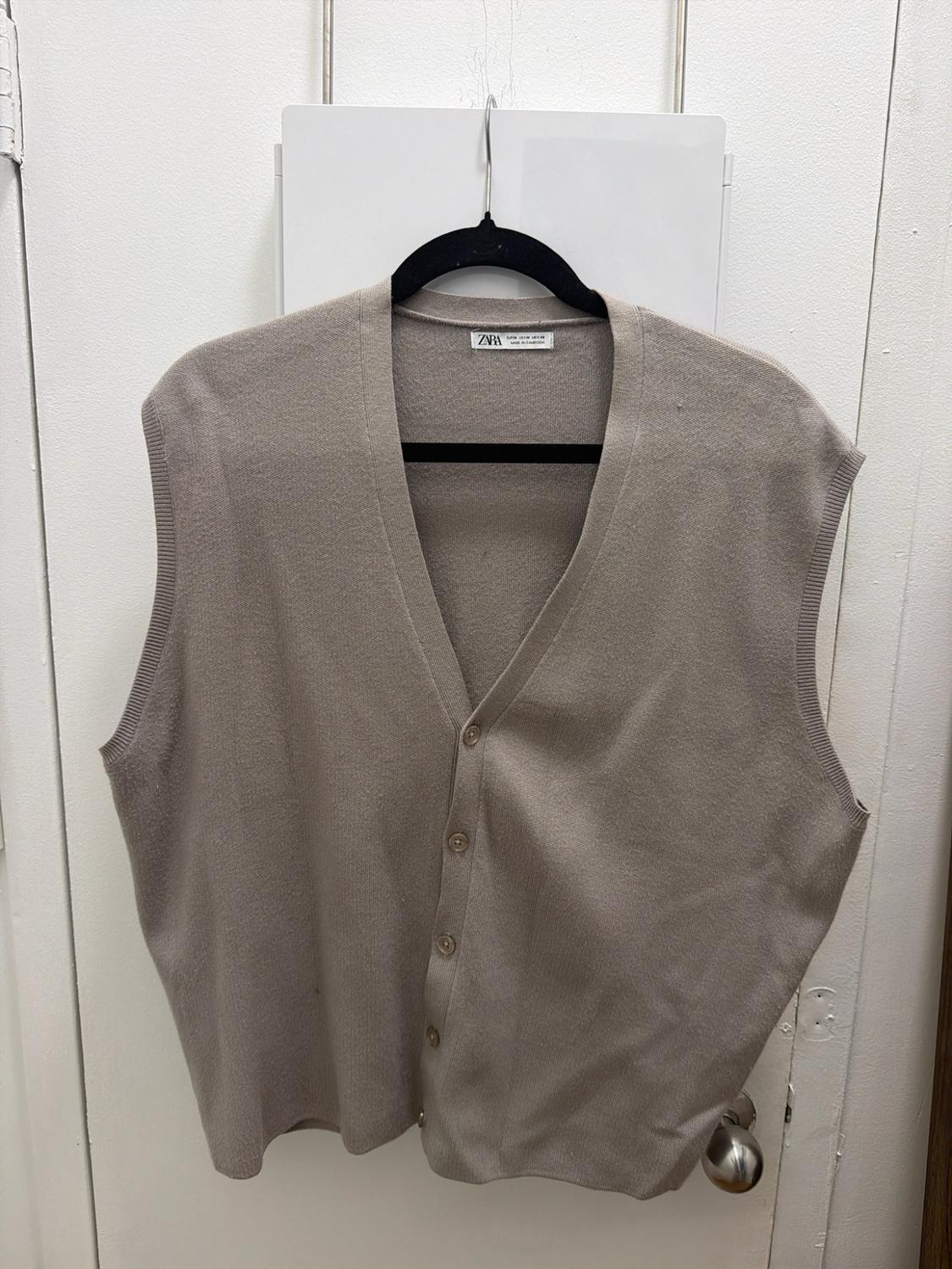 Beige knit vest with a V-neck and front buttons, hanging on a black hanger on a white door. Tag with ZARA text.