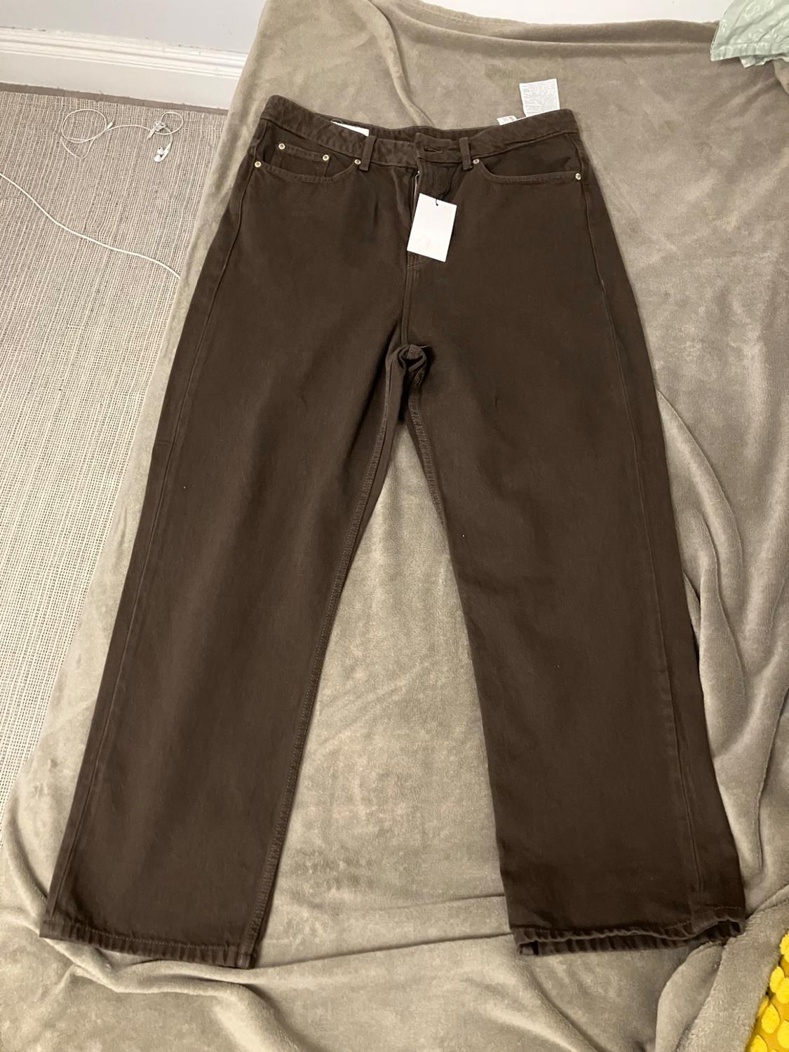 Brown jeans with a tag, laid out on a light-coloured blanket. To the left, there is a cable and white earphones.