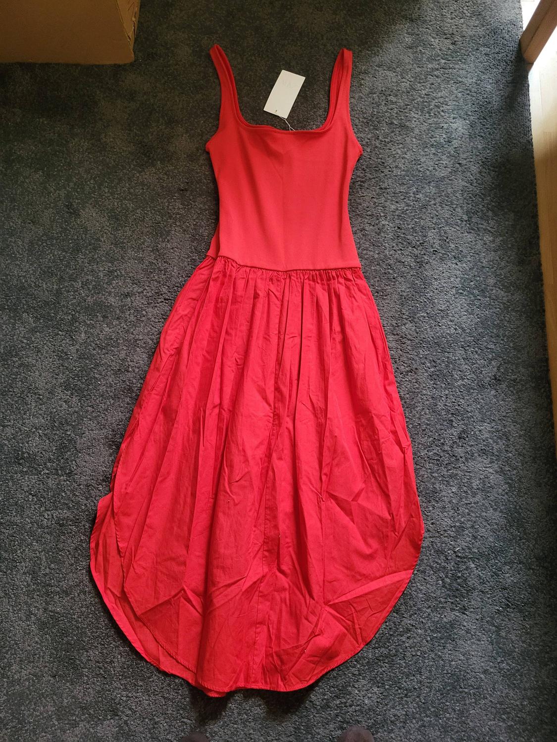 Sleeveless red dress with a full skirt, placed on a grey rug. White label visible at the top.