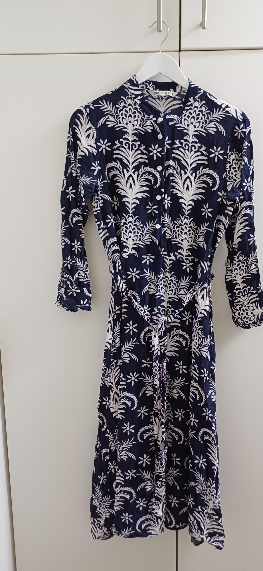 Blue dress with white floral print, long sleeves, hanging on a white hanger in front of a white wardrobe.