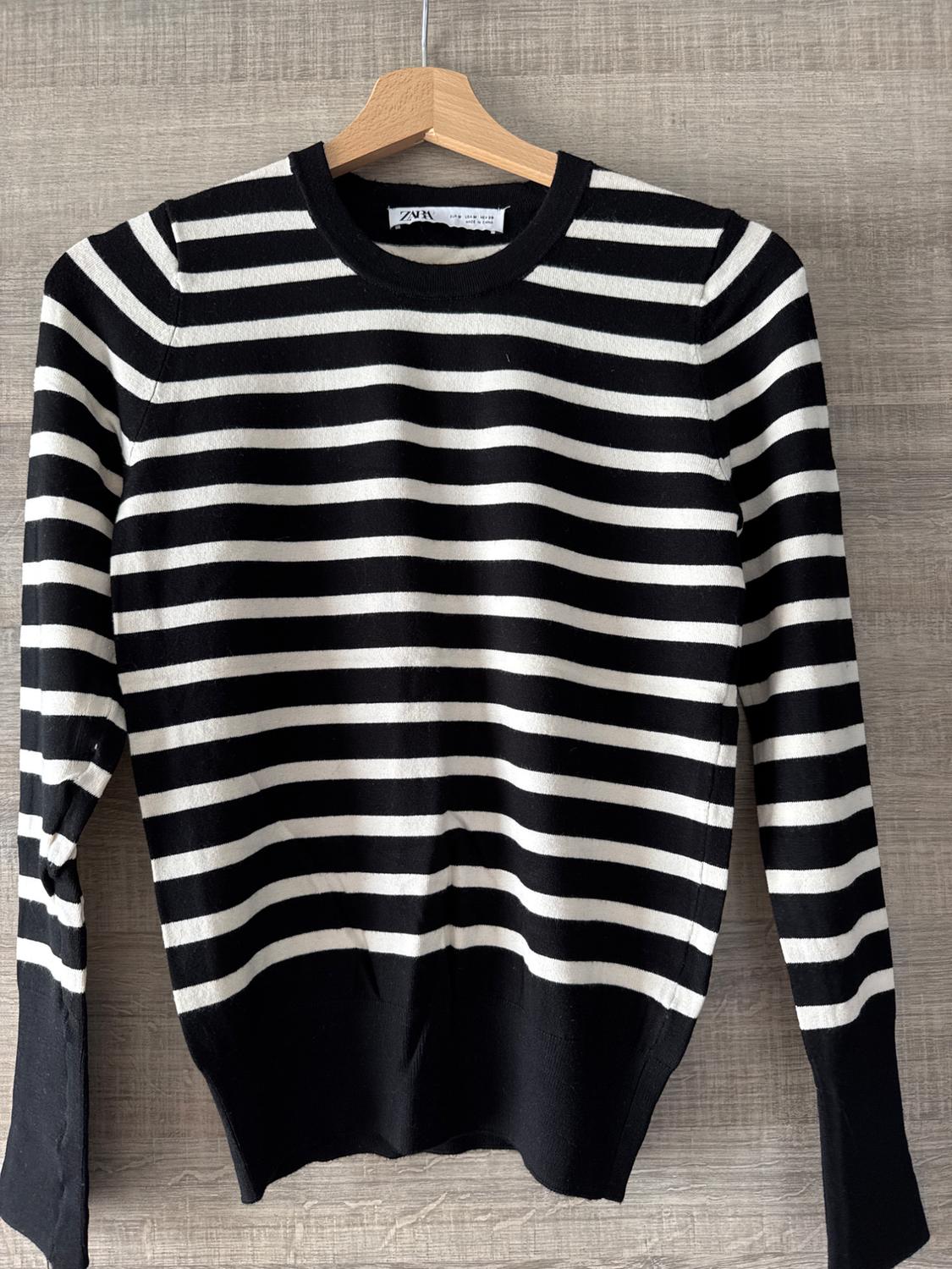 Black and white striped jumper hanging on a wooden hanger against a textured wall background. Label visible.