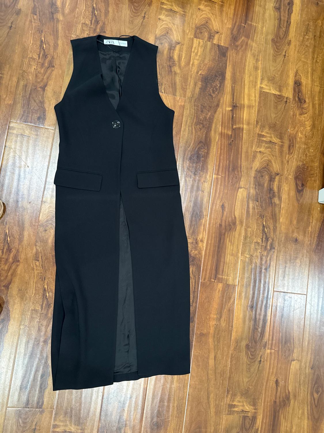 Long black sleeveless waistcoat with lapels and one button, on a brown wooden floor. Label visible on the inside.