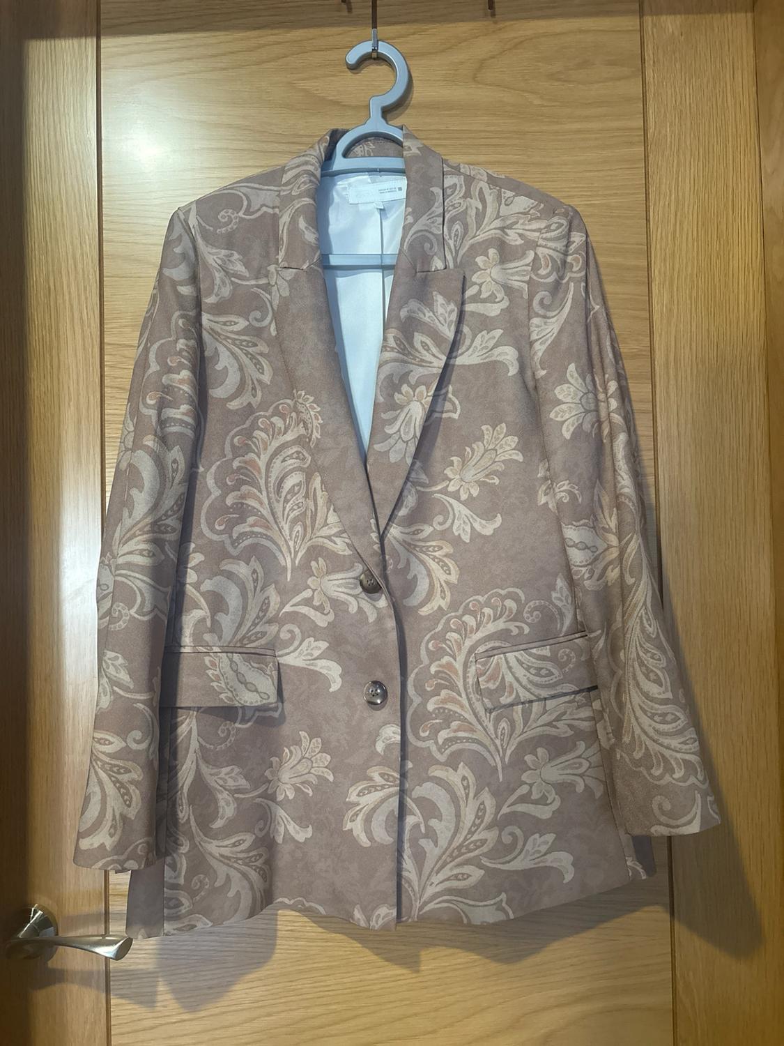 Beige blazer with a light floral print, hanging on a blue hanger on a wooden door.