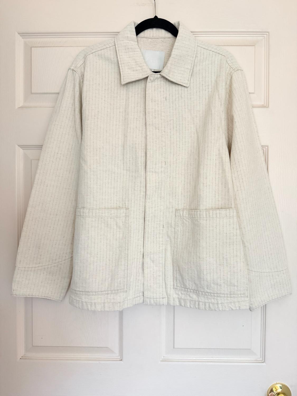 Light-colored jacket with thin vertical stripes and front pockets, hanging on a black hanger over a white door.