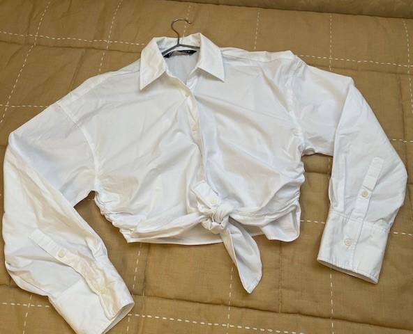 White long-sleeved shirt with a front knot, placed on a beige quilt with visible stitching.