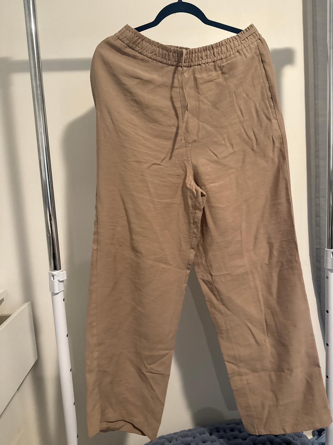 Beige trousers hanging on a black hanger against a light background. The fabric appears light and comfortable, with an elasticated waist.