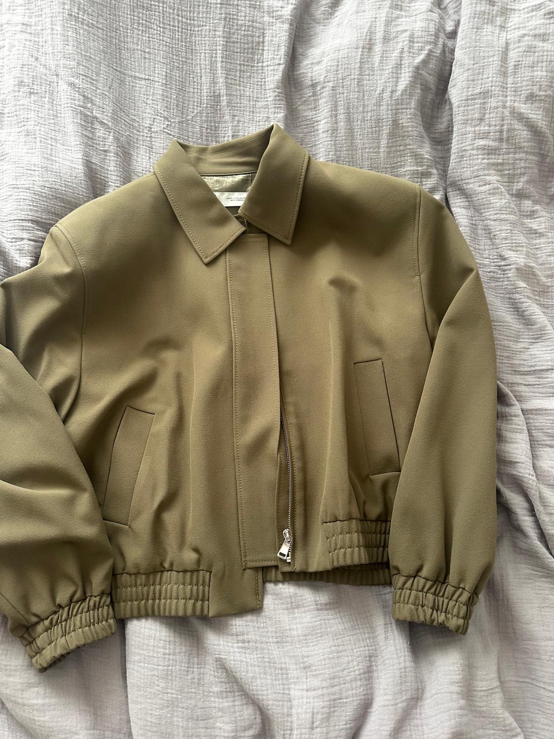Olive green, short jacket with a zip and pockets, over a light grey crinkled fabric.
