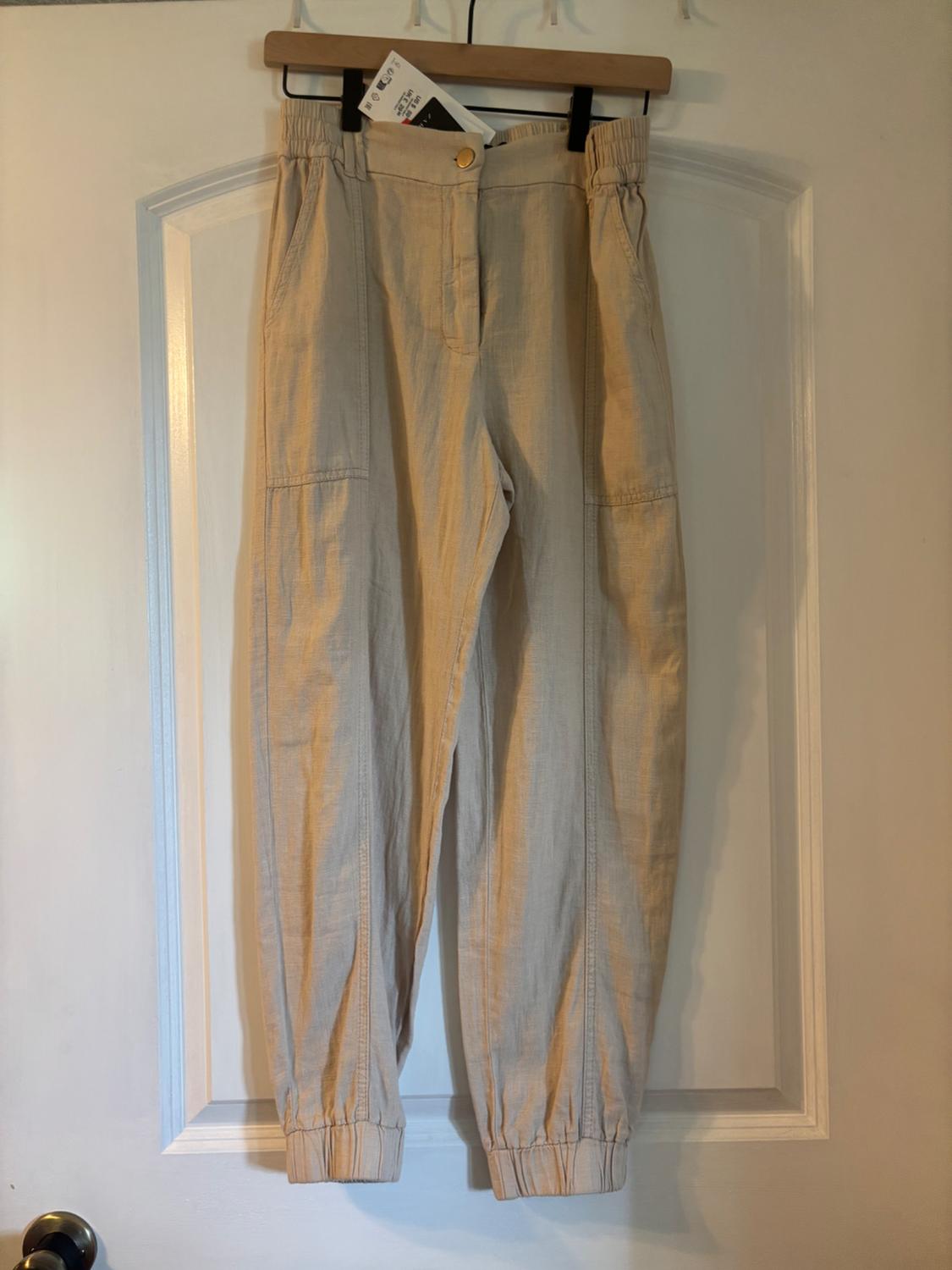 Beige casual trousers with an elasticated waist and cuffs, hanging on a wooden hanger on a white door. Relaxed style.