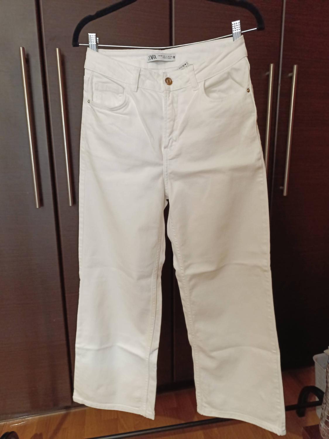 White trousers hanging on a hanger in front of a dark wardrobe. Label visible on the waistband.