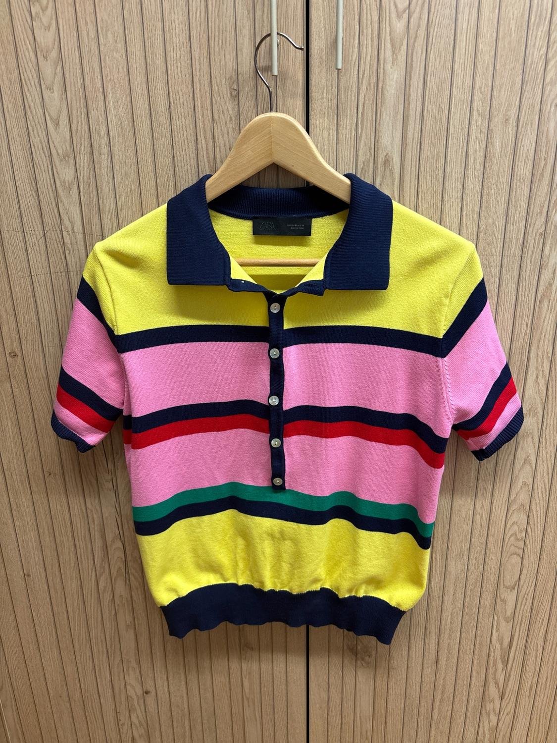 Short-sleeved knitted polo shirt with yellow, pink, red, green and navy blue stripes. Buttoned collar. Hanging on wood. Text: ZARA.