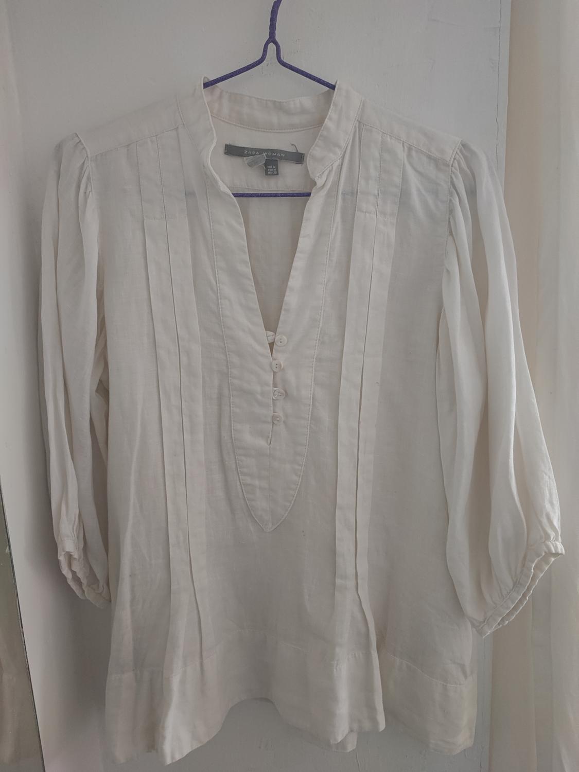 White long-sleeved blouse with pleats and buttons, hanging on a blue hanger. Visible label: "Zara Woman". White background.
