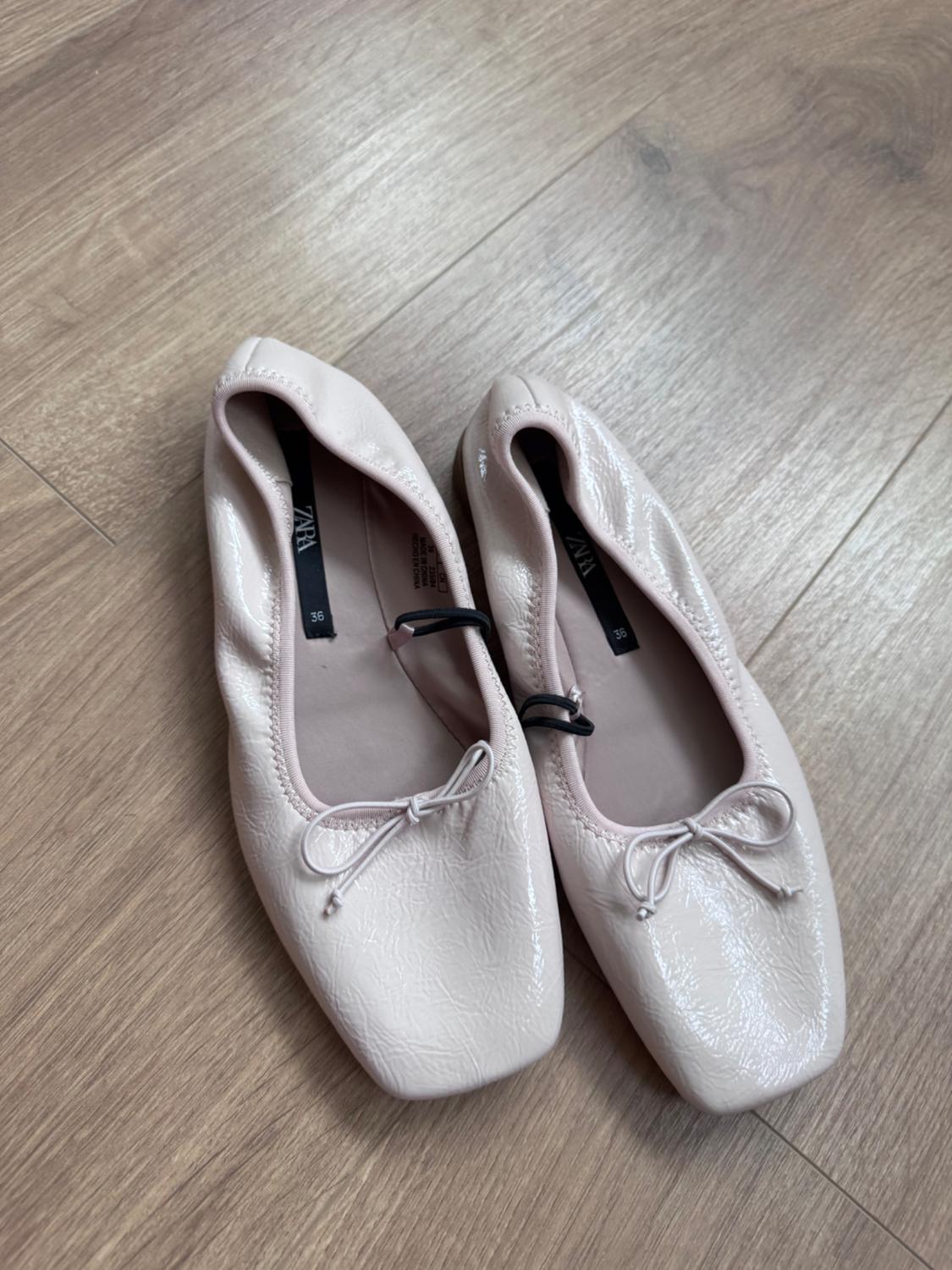 Pair of pink ballet flats with a glossy finish, a bow and square toe on a wooden floor. Visible text: ZARA 36.