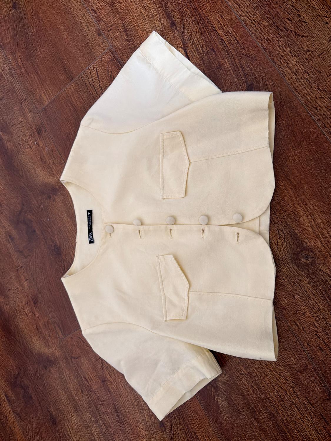 Short beige jacket with buttons and front pockets, on a brown wooden floor. Visible label on the collar.