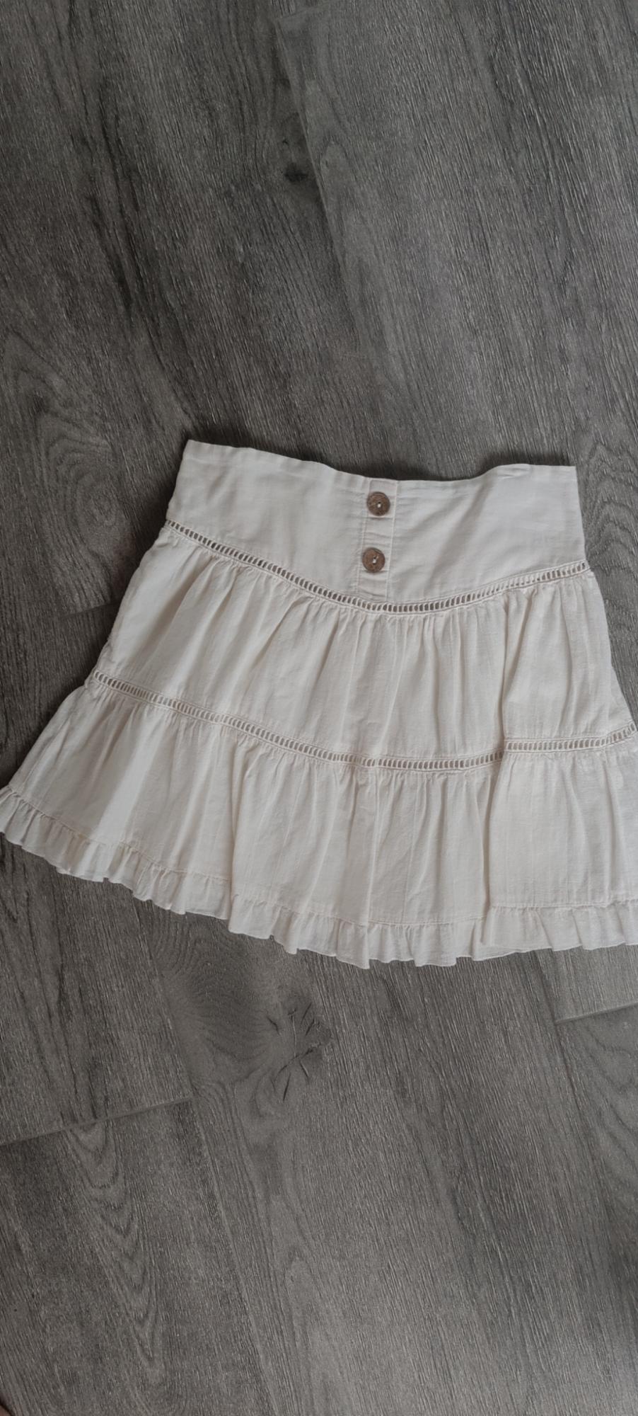 White, casual-style short skirt with ruffles, openwork details, and two buttons, on a grey wooden floor.