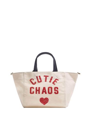Beige fabric bag with black handles. Red text "CUTIE CHAOS" and a heart in the center. White background.