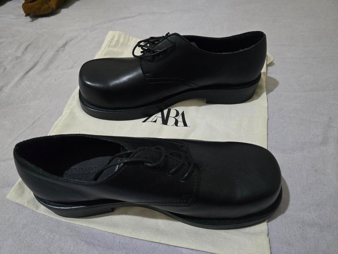 Black round-toe lace-up shoes on a fabric bag bearing the text ZARA, resting on a grey fabric surface.