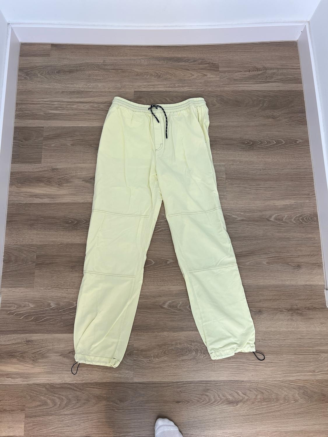 Light yellow trousers with an adjustable drawstring and knee seams, on a wooden floor. One foot is visible at the bottom edge.