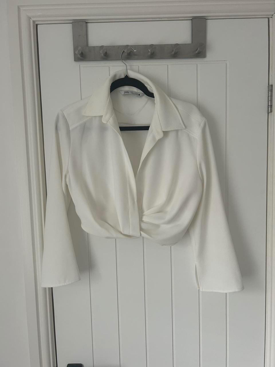 Long-sleeved white blouse with a wrap-around design, hung on a white door. Visible tag: "ZARA".