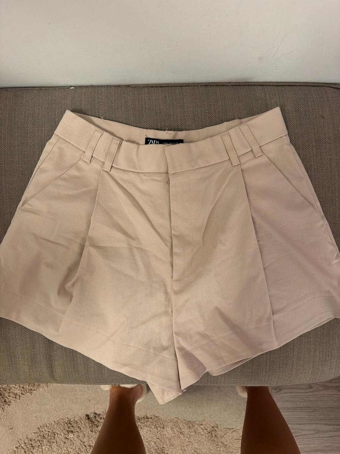 Μπεζ σορτς με πιέτες και ετικέτα ZARA πάνω σε μια γκρι επιφάνεια. Στο κάτω μέρος φαίνονται τα πόδια ενός ατόμου.