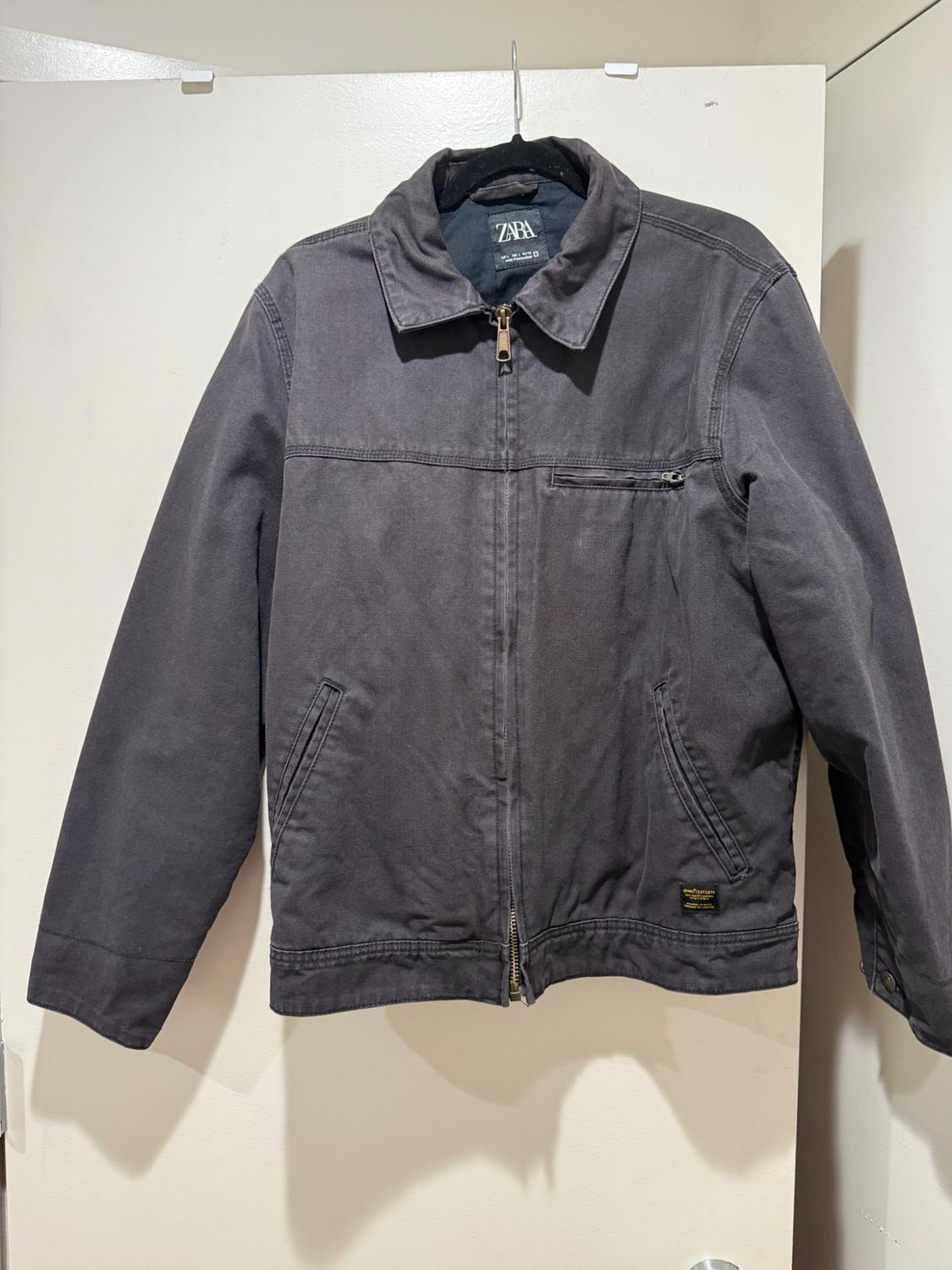 Dark gray jacket with a zipper, collar and pockets, hanging on a hanger over a white door. A Zara brand label is visible.