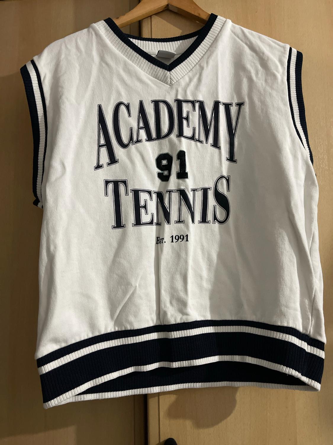 White V-neck vest with navy blue stripes. Text: ACADEMY 91 TENNIS Est. 1991. Hanging on a wooden coat hanger.