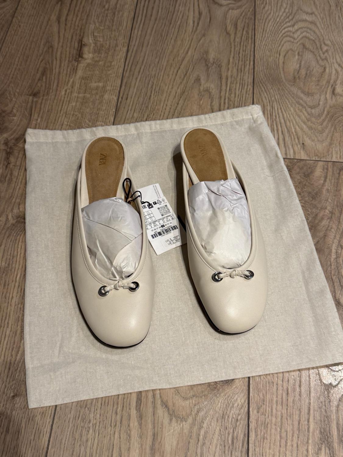 Cream slingback shoes with a bow on a fabric bag against a wooden floor. They are stuffed with paper and have a tag. Text: ZARA.