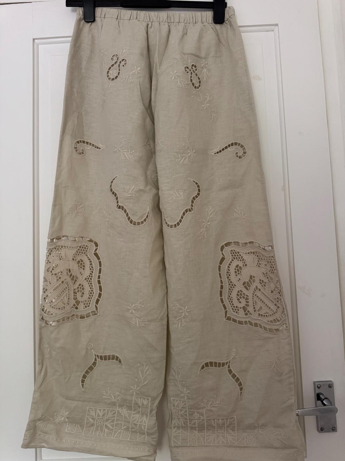Beige wide-leg trousers in a relaxed style with an elasticated waist and broderie anglaise detailing, hanging on a black hanger against a white door.