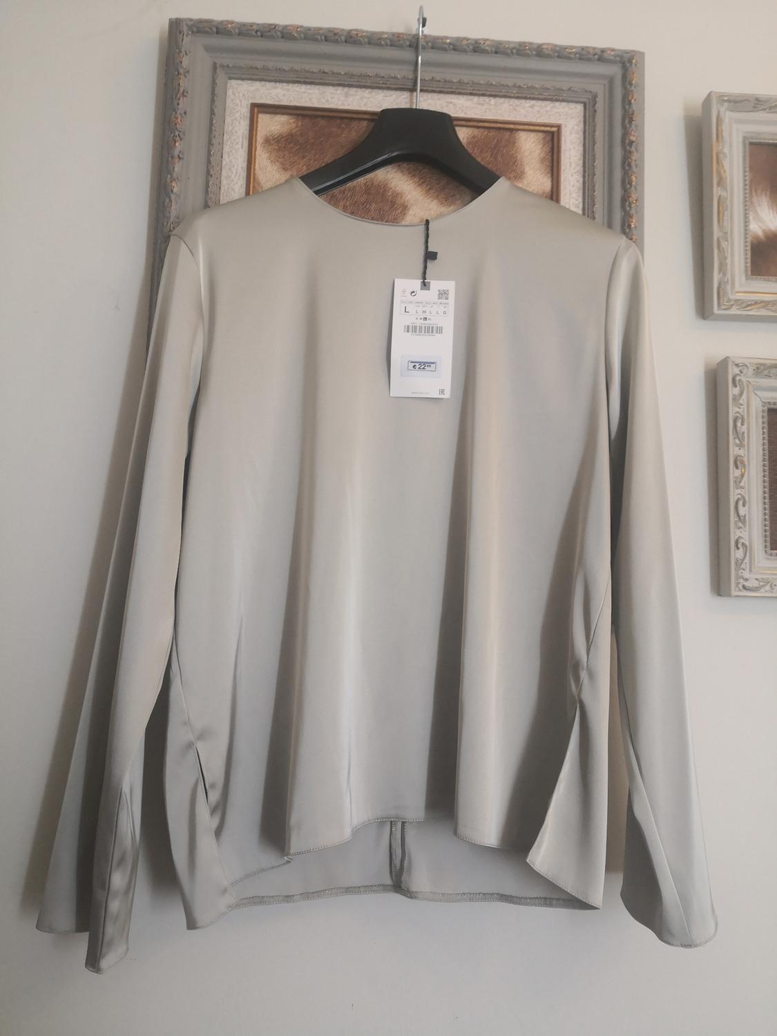 Beige, long-sleeved blouse on a black hanger, with visible label. Decorative background with frames and textures in neutral tones.